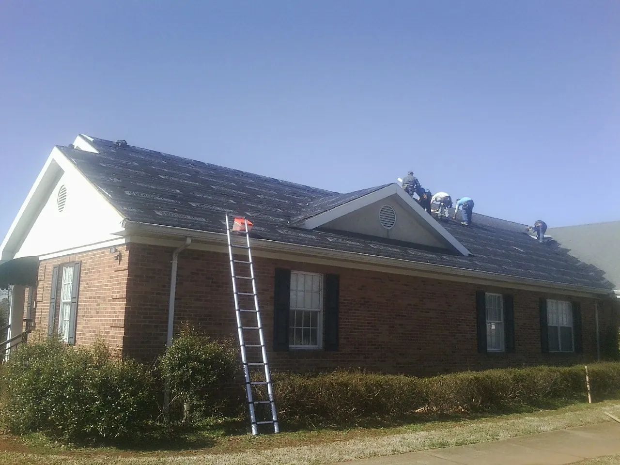 Crew tearing shingles off a brick ranch roof with a ladder set — Metal Roof Repair work by our crew in Cumbola
