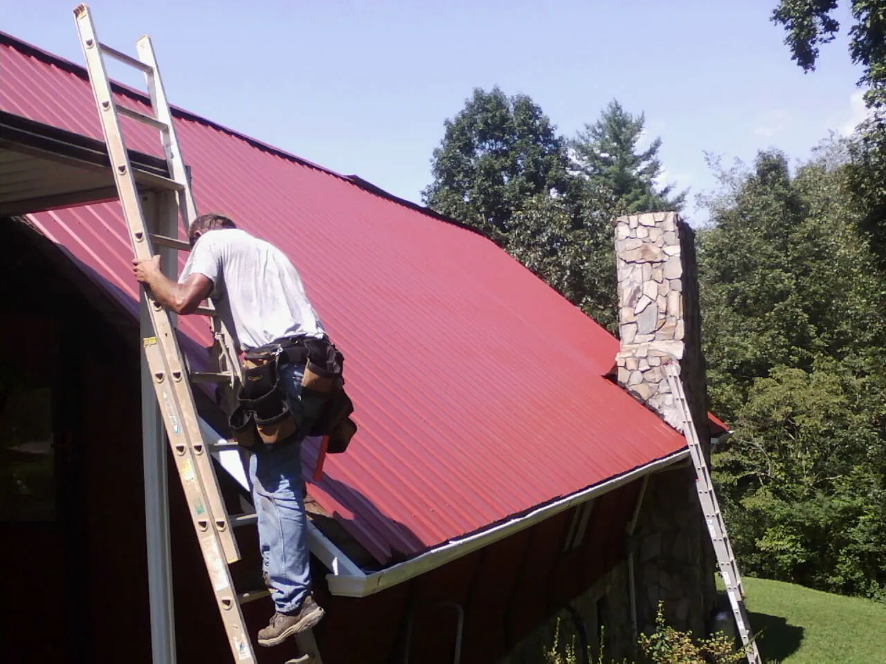 Roofer on a ladder beside a red metal roof and stone chimney — Soffit & Fascia Repair by Roofing Brothers in Cumbola